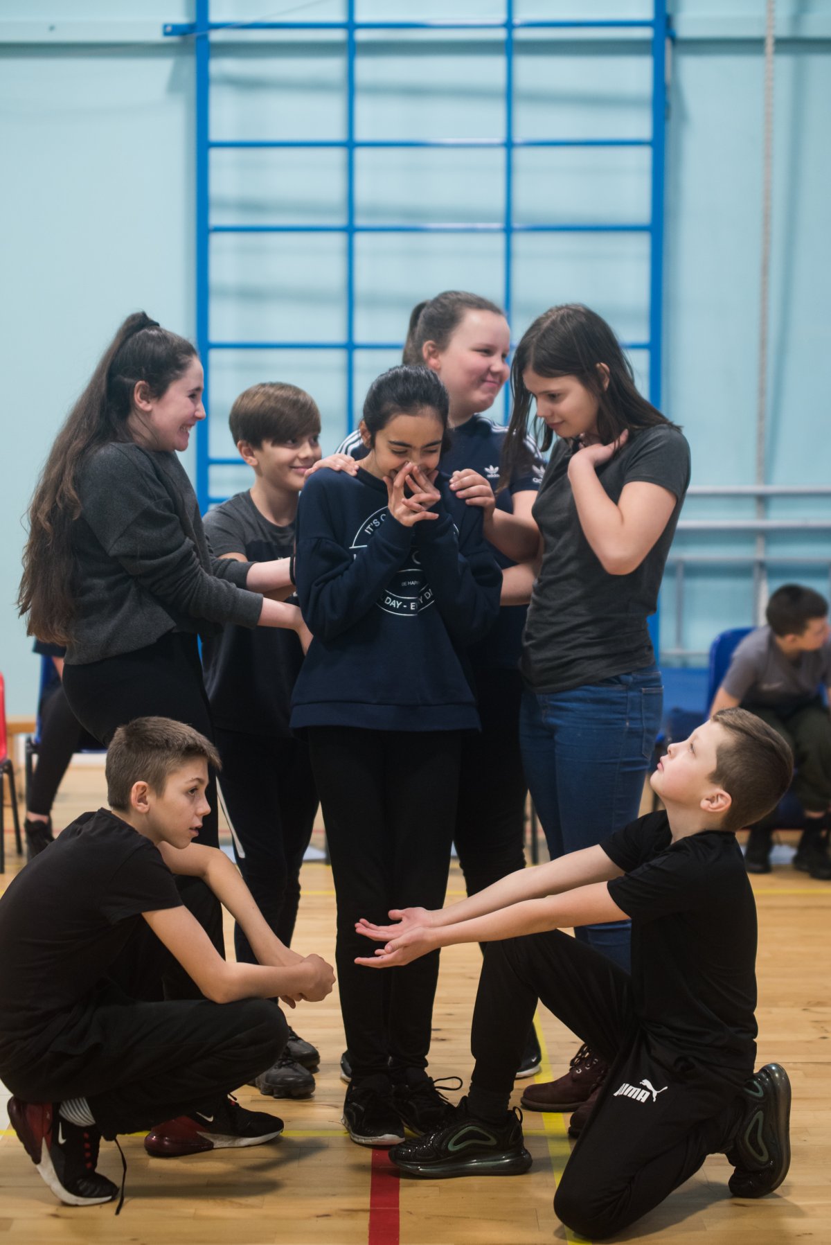 A group of children are in a school hall taking part in a movement activity. They are smiling and encouraging the girl at the centre of the group to take a step of faith and trust the other members of the group that she will be safe.