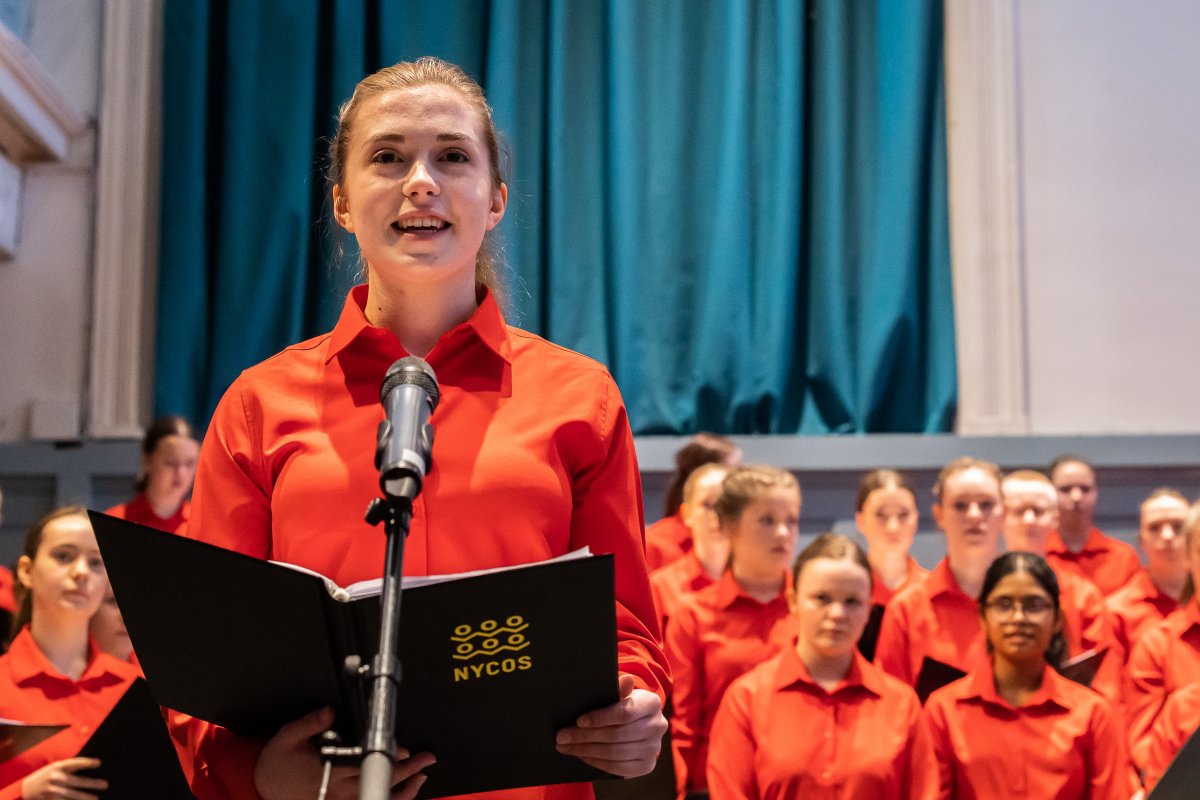 A teenage girl stands in front of a microphone singing. Behind her are other girls in the choir. They are all wearing orange shirts and holding folders with 'NYCOS' on the front.