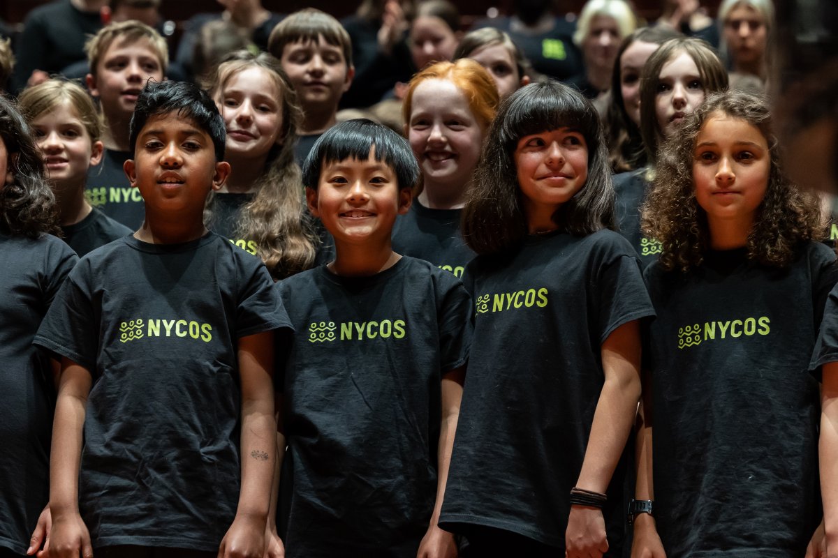 Primary school aged children are smiling and singing as a choir. They are wearing black t-shirts with "NYCOS" on the front. The children represent a diverse array of ethnic backgrounds.